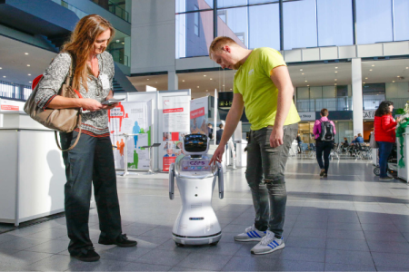 Ein Mann (rechts) und eine Frau (link) stehen an einem Mesestand und schauen sich einen Pflegeroboter an. Der Mann führt der Frau den Roboter vor und zeigt verschiedene Funktionen.