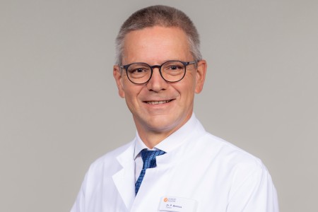 You can see a portrait photo of Dr. Peter Bernius, chief physician at the Schön-Klinik's Centre for Paediatric and Neuro-Orthopaedics. He is wearing a white doctor's coat and a tie. He is also wearing glasses, has short grey hair and is smiling at the camera.