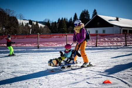 Eine Frau steht auf zwei Skiern. Sie schiebt ein Kind an. Dieses sitzt, unter ihm sind Skier.