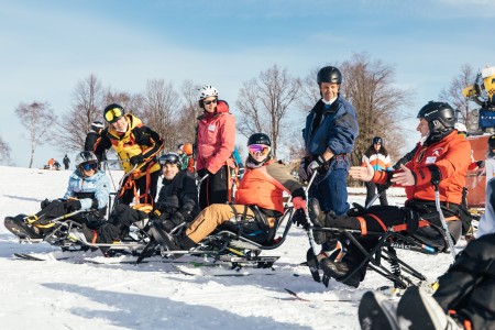 Seven people are on skis. Four of them are sitting on ski equipment for people with disabilities, three people are standing on skis. Six people are looking at a man who is also sitting on skis. He is explaining how to handle the skis.