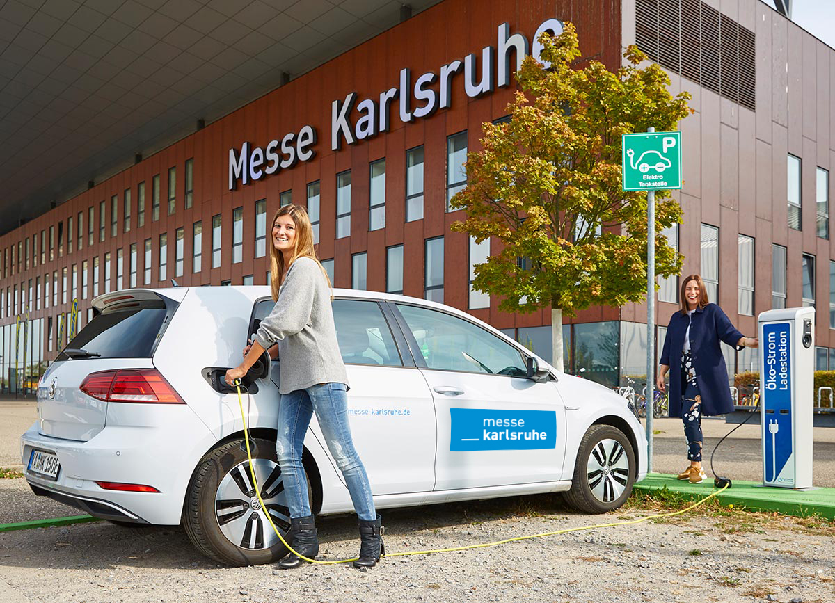 Foto der Ladesäule für Elektrofahrzeuge vor der Messe Karlsruhe. Zwei Frauen laden ein Elektrofahrzeug der Messe Karlsruhe damit.