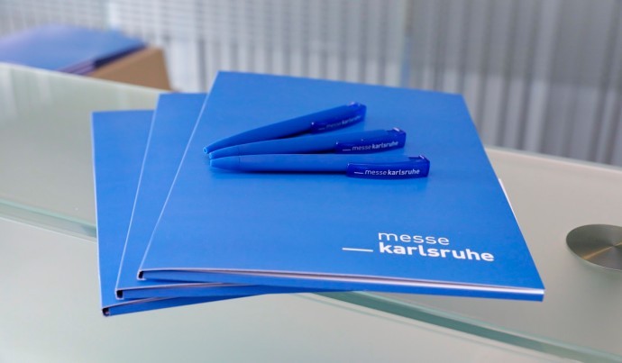 Photo of three blue press kits stacked on top of each other and three blue pens with the logo of Messe Karlsruhe. 