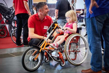 Young girl sits in a wheelchair with three wheels and talks to an exhibitor at REHAB 