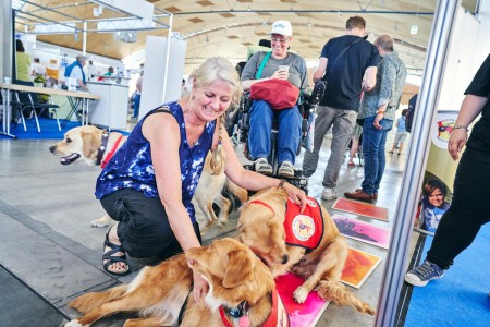 Woman stroking two self-help dogs. A person in a wheelchair sits next to them and smiles.