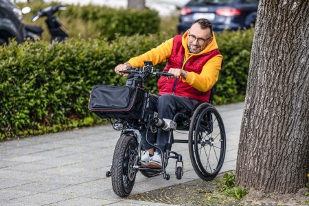 Foto von einem Mann, der im Rollstuhl mit PAWS-Zuggerät eine Straße entlang fährt.