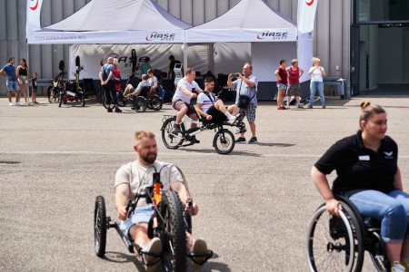 In the foreground, people are testing out various riding devices such as handbikes, bicycles and wheelchairs. In the background is a stand with a large selection of different prdoucts for active wheelchair users.