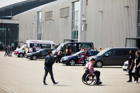 Numerous cars can be seen parked along the exhibition hall and inviting people to test drive them. In the foreground are people walking and in wheelchairs on their way to the next hall.