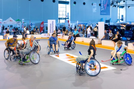  Neun Erwachsene und Kinder in Rollstühlen spielen in der Inklusiven Sportstätte mit Begeisterung Hockey.